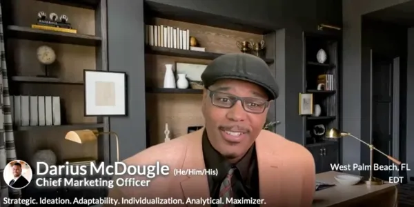 Dr. Darius McDougle, Chief Marketing Officer, speaking on camera in a peach blazer and black cap